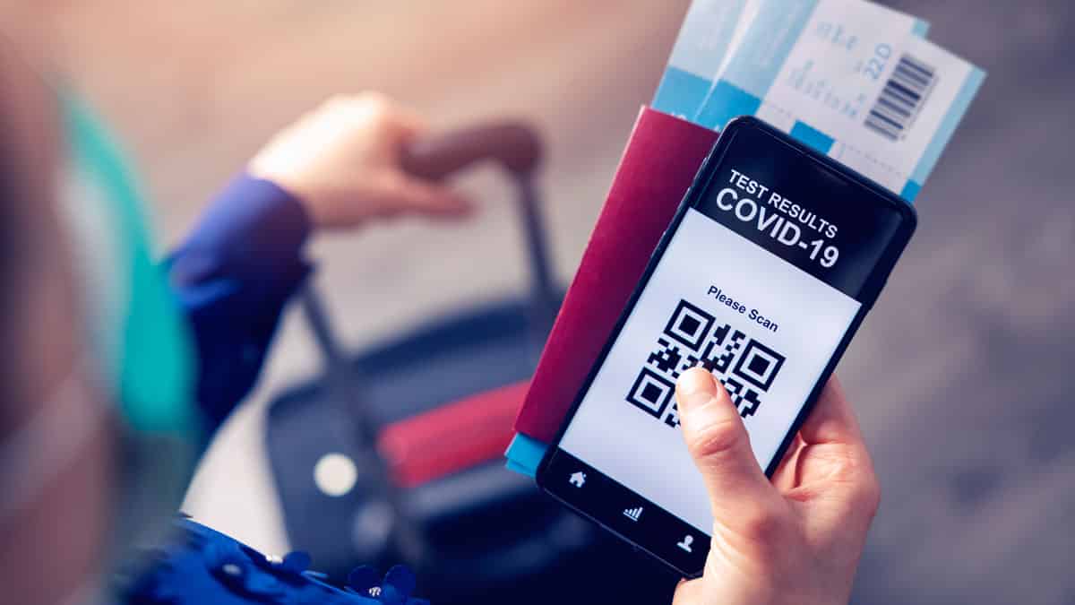 Person med koffert i den ene hånden, pass, flybiletter og digital covid-sertifikat på mobilen i den andre hånden