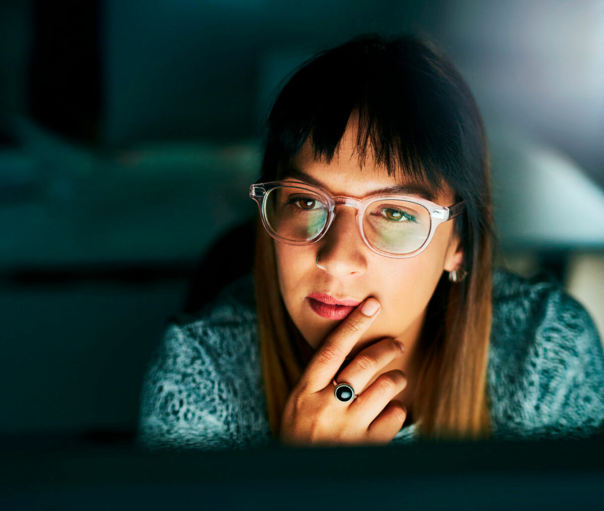 ung kvinne med langt hår og briller sitter i mørket og ser på opplyst dataskjerm