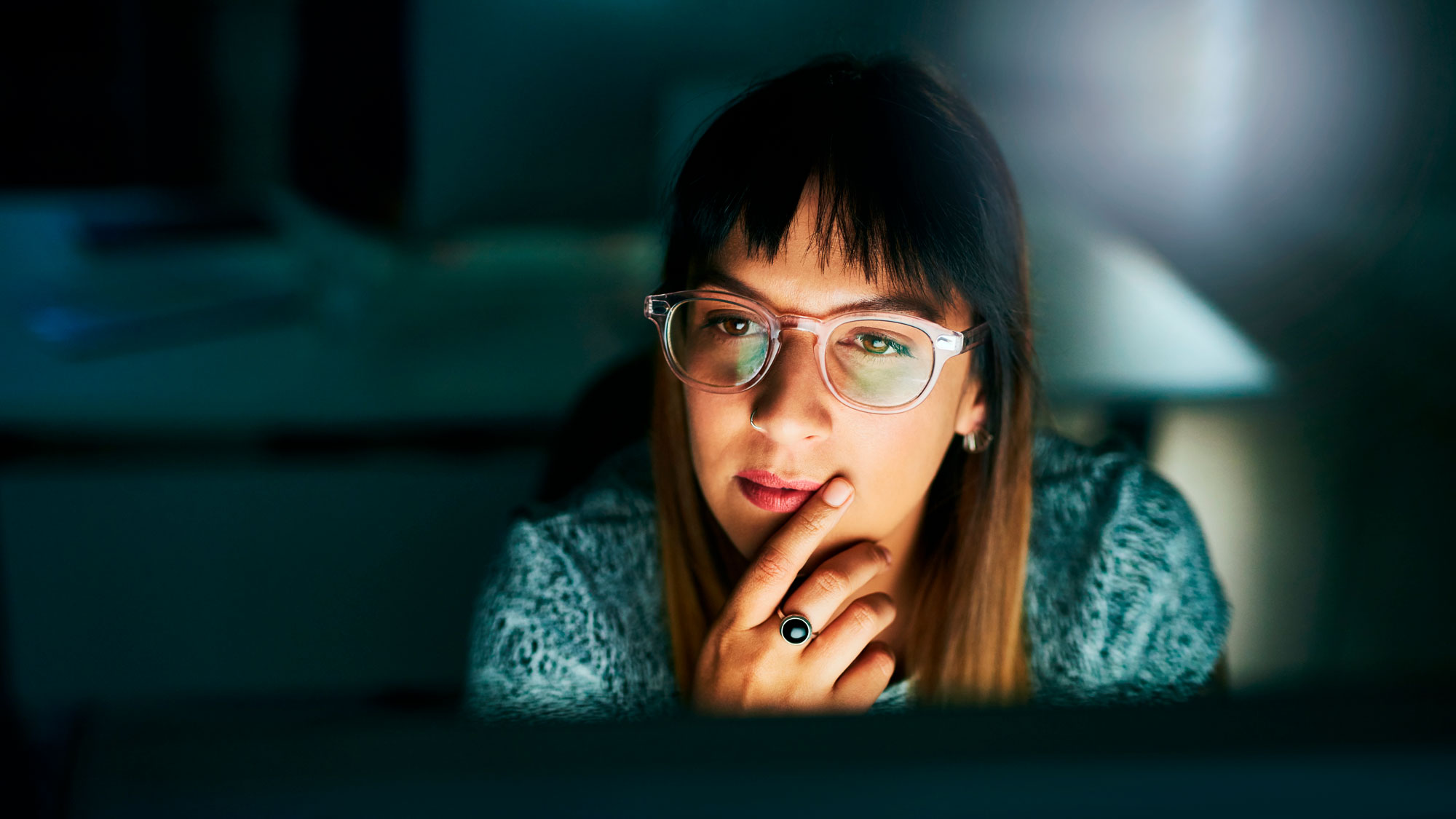 ung kvinne med langt hår og briller sitter i mørket og ser på opplyst dataskjerm